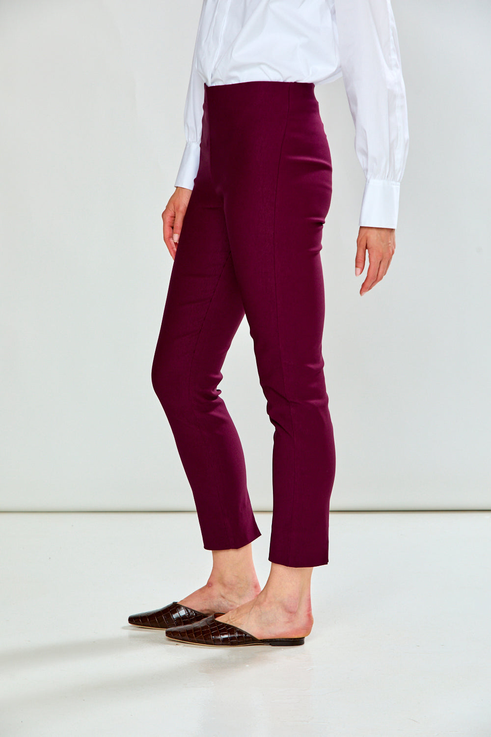 A person wearing high-waisted, ankle-length burgundy pants, styled with a white long-sleeve blouse and dark brown mule flats, shown from the waist down against a plain white studio background.