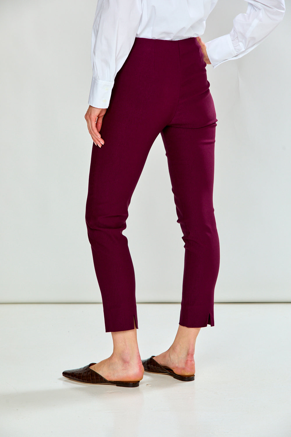 A person wearing high-waisted, cropped ankle-length pants in deep burgundy, paired with a white long-sleeve blouse and dark brown mule flats, shown from a side-back angle against a plain white studio background.