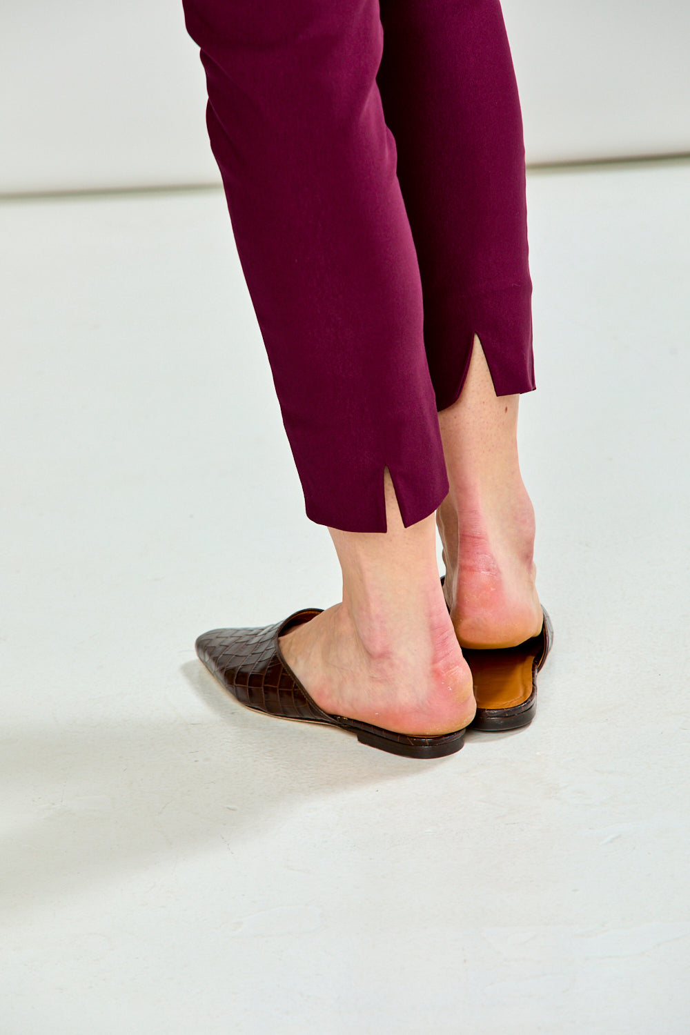 Close-up of a person's feet wearing dark brown pointed-toe mule flats and deep burgundy pants, standing on a light gray studio floor.