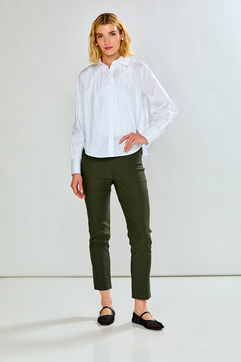 A model wearing green cropped trousers, styled with a white long-sleeve button-up shirt and black flat shoes, shown from the waist down against a plain white studio background.