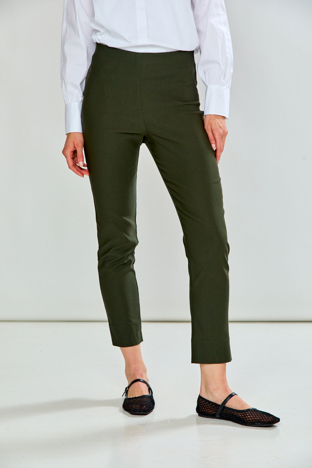 A model wearing green cropped trousers, styled with a white long-sleeve button-up shirt and black flat shoes, shown from the waist down against a plain white studio background.