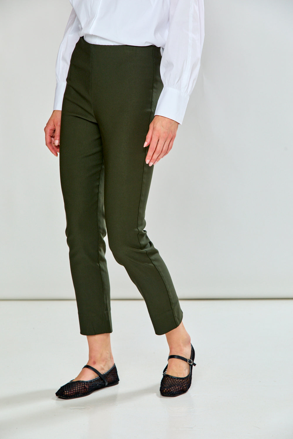 A model wearing green cropped trousers, styled with a white long-sleeve button-up shirt and black flat shoes, shown from the waist down against a plain white studio background.