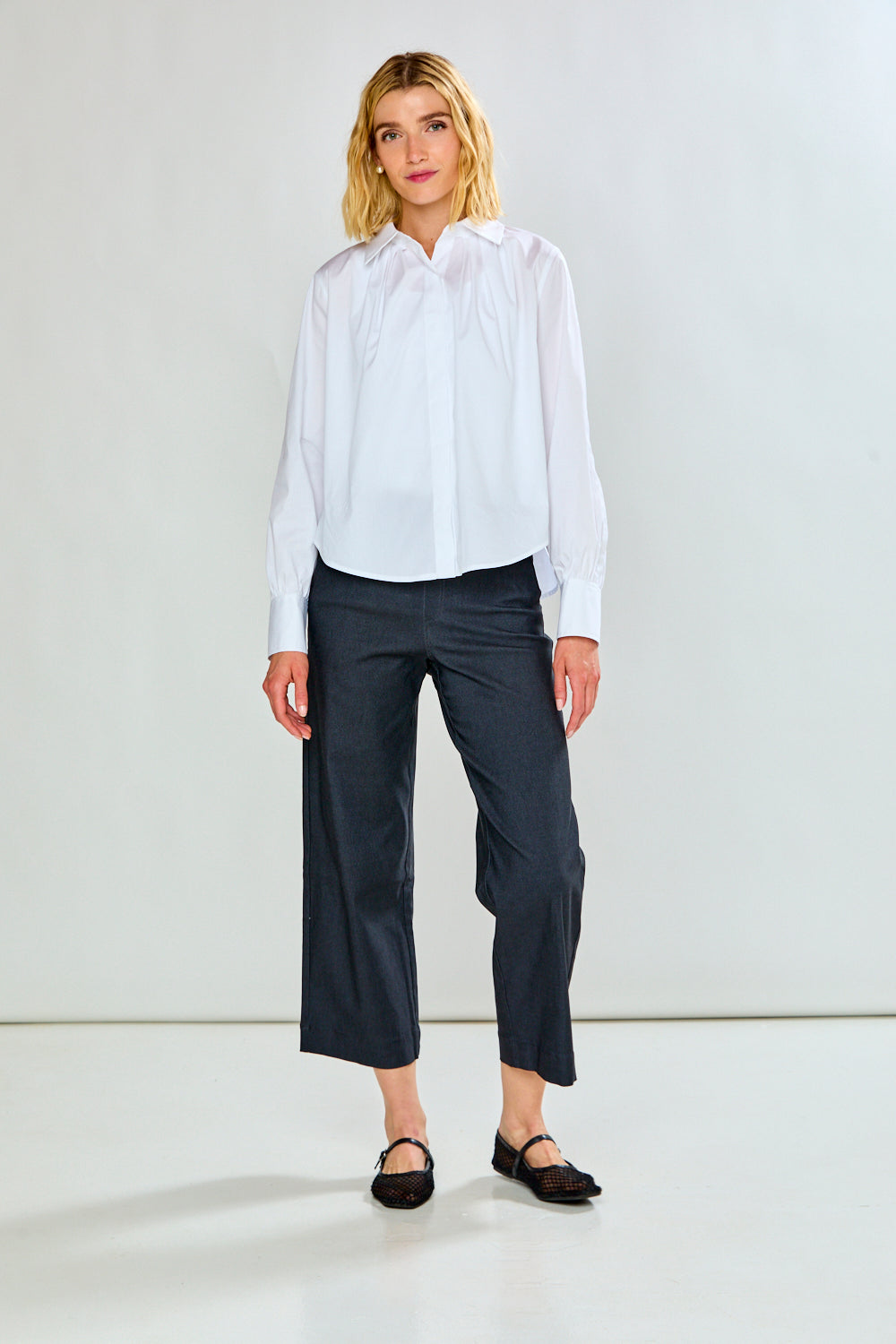 A model wearing charcoal cropped trousers with a white long-sleeve shirt and black flat shoes, standing against a light gray studio background.