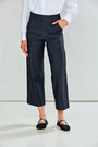 A model wearing charcoal cropped trousers with a white long-sleeve shirt and black flat shoes, standing against a light gray studio background.