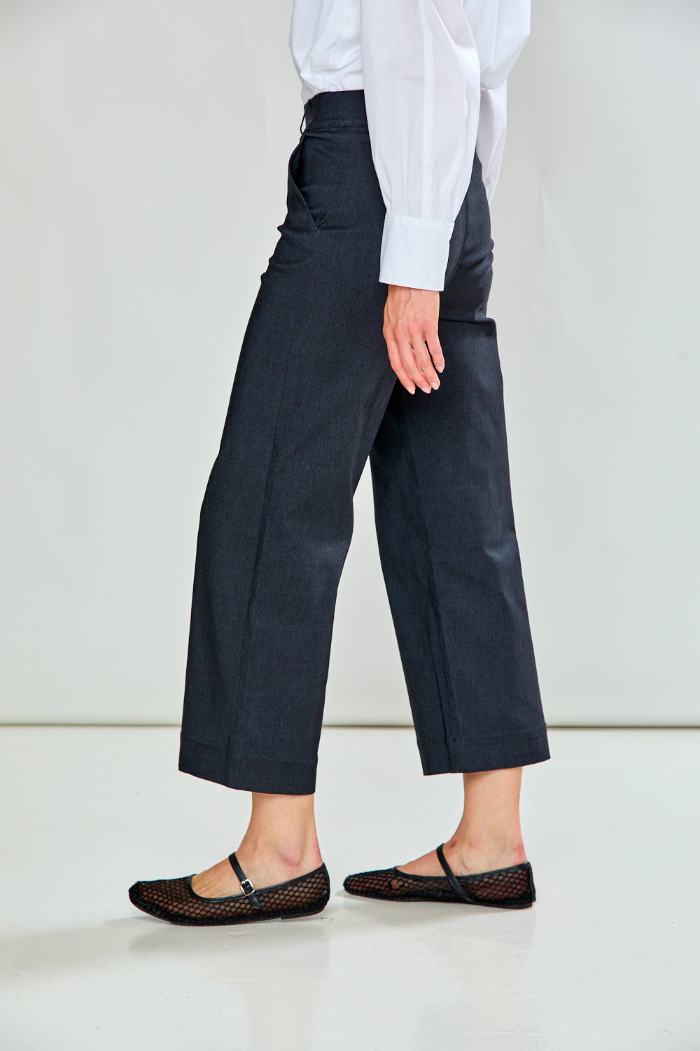 A model wearing charcoal cropped trousers with a white long-sleeve shirt and black flat shoes, standing against a light gray studio background.