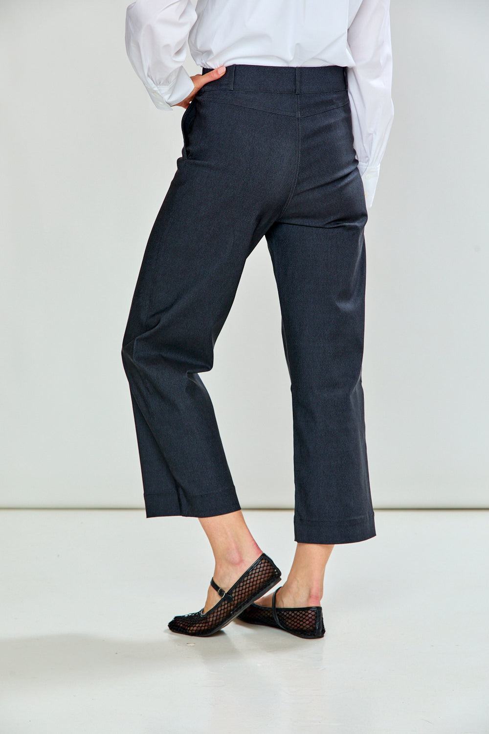 A model wearing charcoal cropped trousers with a white long-sleeve shirt and black flat shoes, standing against a light gray studio background.