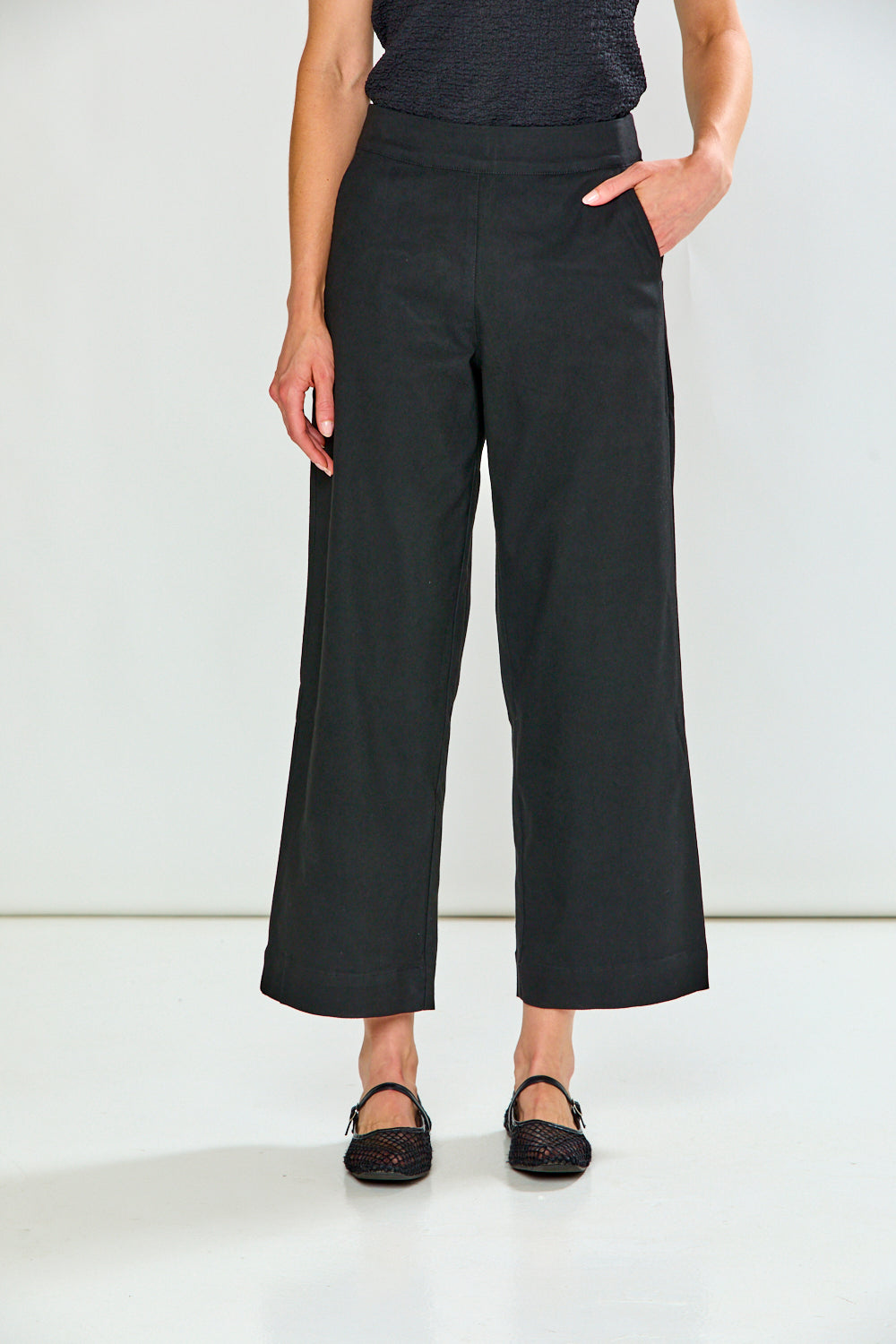 A model wearing black wide-leg cropped pants, paired with a dark textured top and black lace-up flats, standing against a clean white studio background.