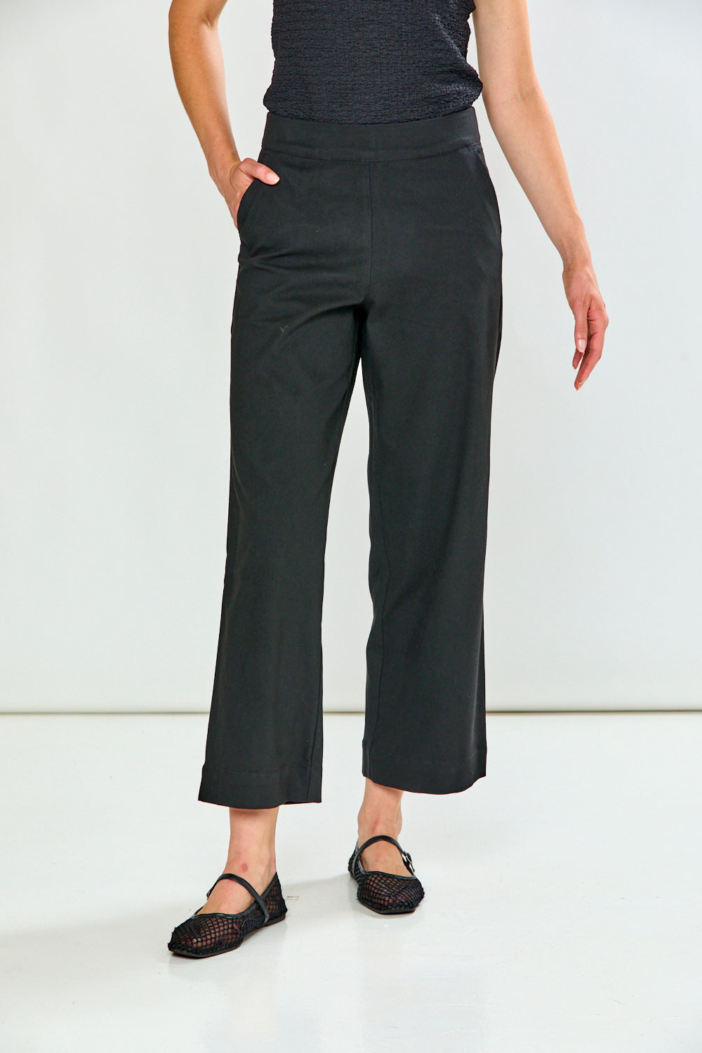 A model wearing black cropped wide-leg pants with side pockets, paired with a dark textured top and black flat shoes, standing against a plain white studio background.