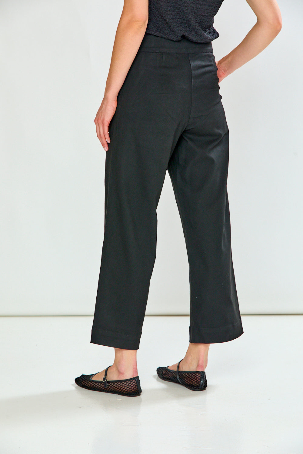 A model from the back wearing cropped trousers, a dark gray top, and black flat shoes with a fishnet pattern, set against a plain white studio background.