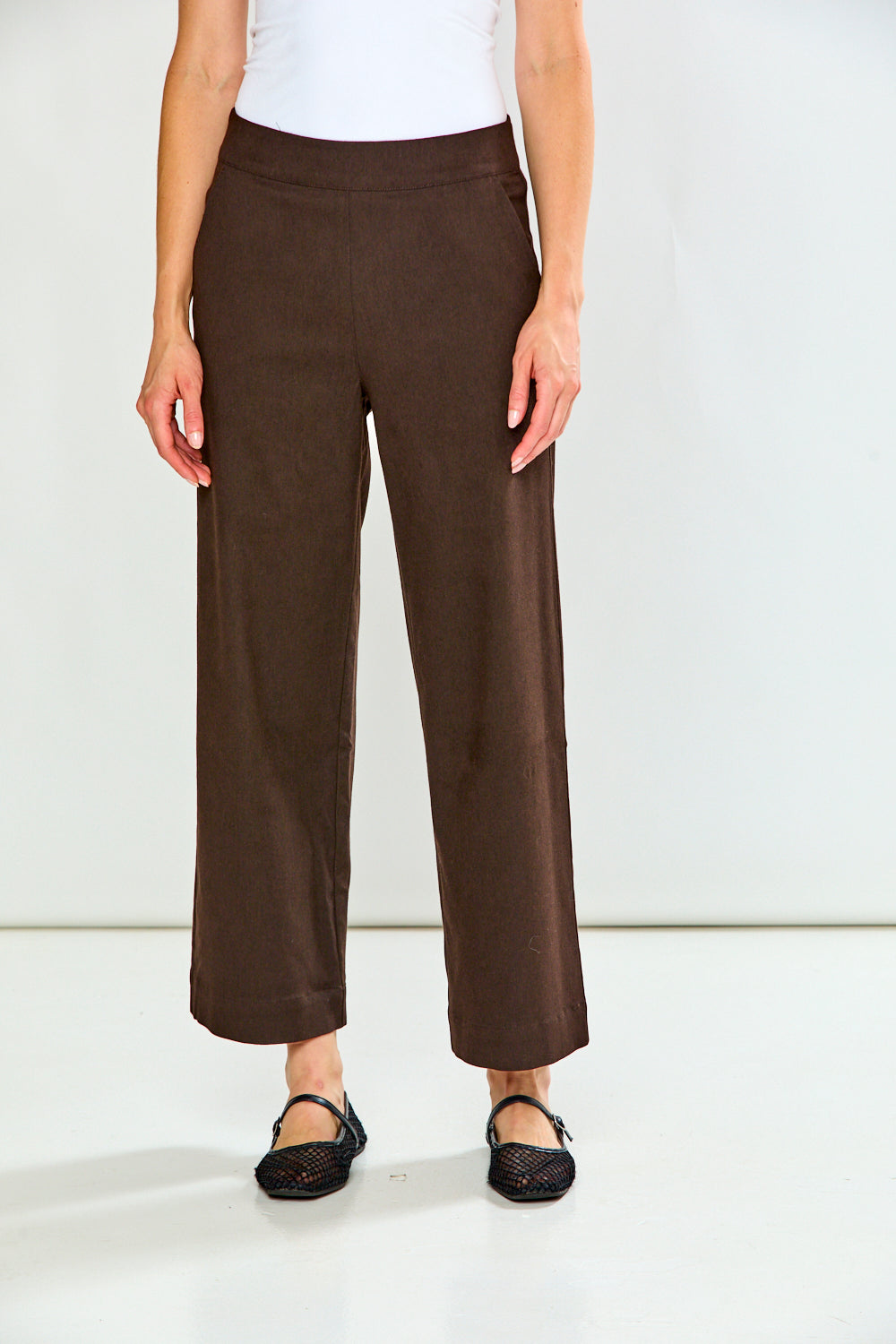 A model wearing brown wide-leg cropped pants, a white top, and black flat shoes, standing against a plain white studio background.