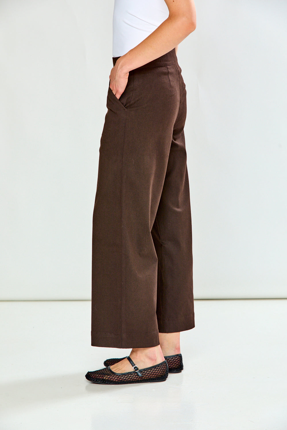 A person shown from the waist down wearing dark brown wide-leg cropped pants, a white top, and black flat shoes against a plain white studio background.