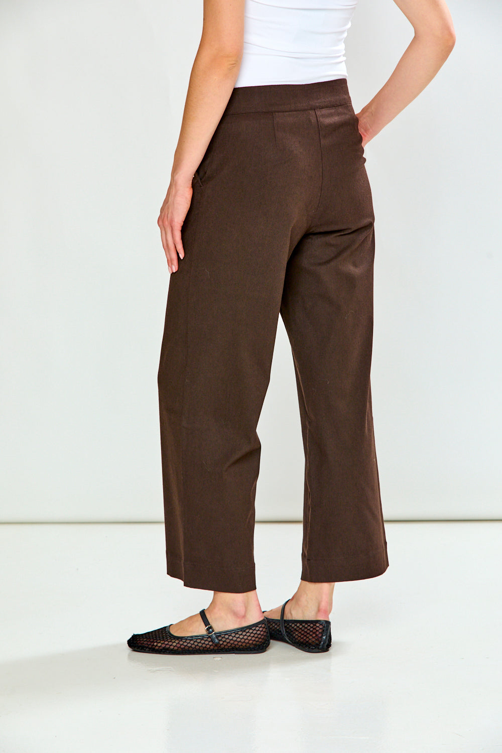 A model from the back wearing brown cropped wide-leg trousers, a white ribbed top, and black flat shoes with a fishnet strap, set against a plain white studio background.