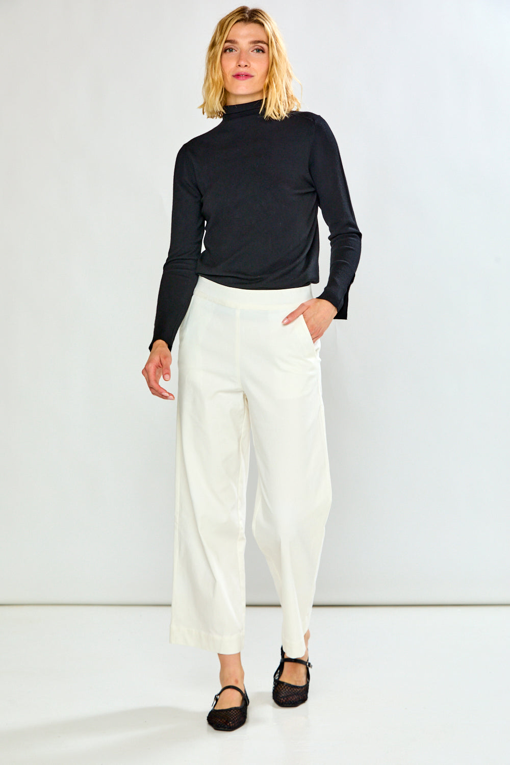 A woman modeling a black long-sleeve mock neck top and white cropped wide-leg trousers with black flat shoes, standing against a light gray studio background.