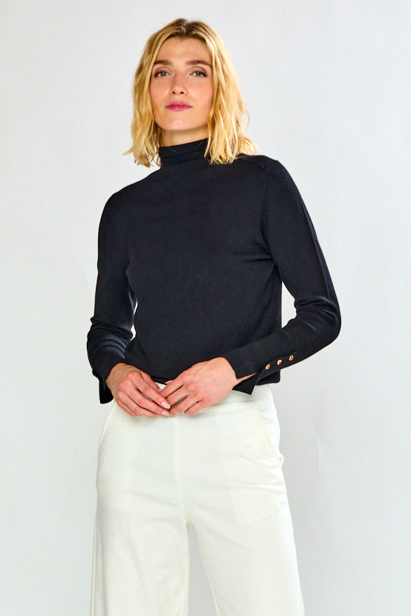 A woman modeling a black long-sleeve mock neck top and white cropped wide-leg trousers, paired with black flat shoes, against a clean white studio background.