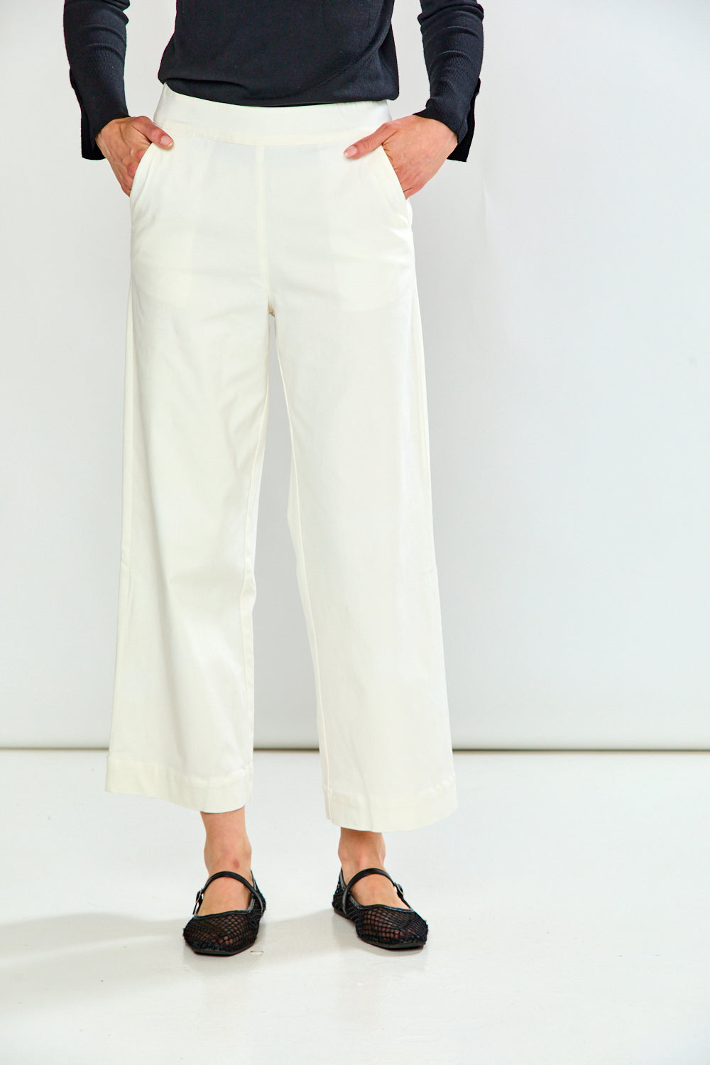 A person wearing off-white cropped wide-leg pants with a black long-sleeve top and black flat shoes, standing against a plain white studio background.