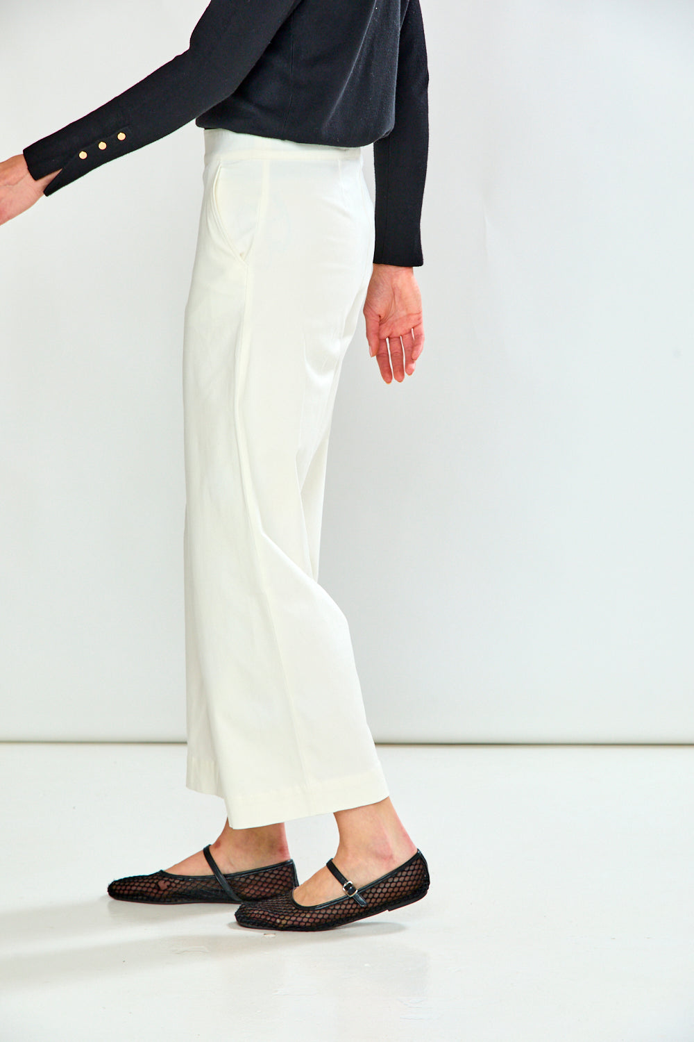 A model is shown from the side wearing a high-waisted, wide-leg white cropped skirt, a black long-sleeve top, and black mesh Mary Jane flats against a clean white studio background.