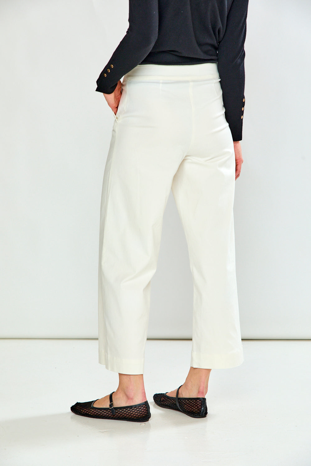 A model shown from the back wearing white cropped trousers, a black long-sleeve top, and black flat shoes against a clean white studio background.