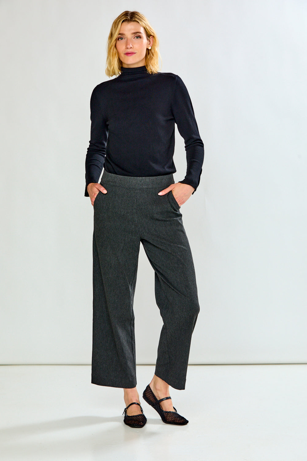 A woman modeling a dark long-sleeve mock neck top and dark gray wide-leg cropped trousers, standing against a plain light gray studio background.