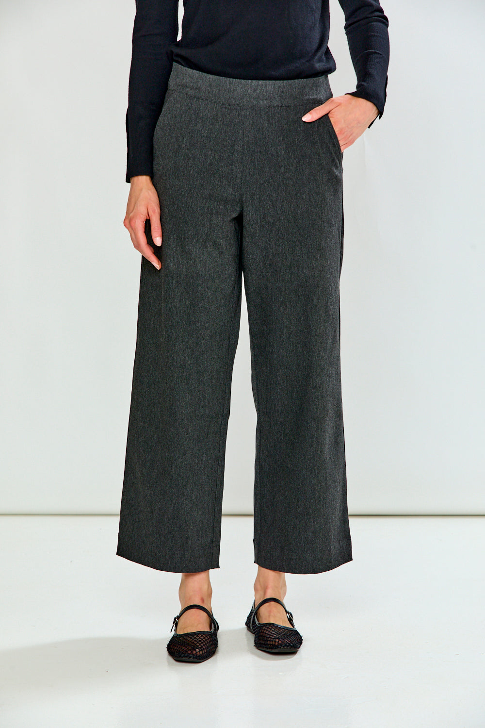 A model wearing dark gray wide-leg cropped trousers, paired with a black long-sleeve top and black flat shoes, standing against a plain white studio background.