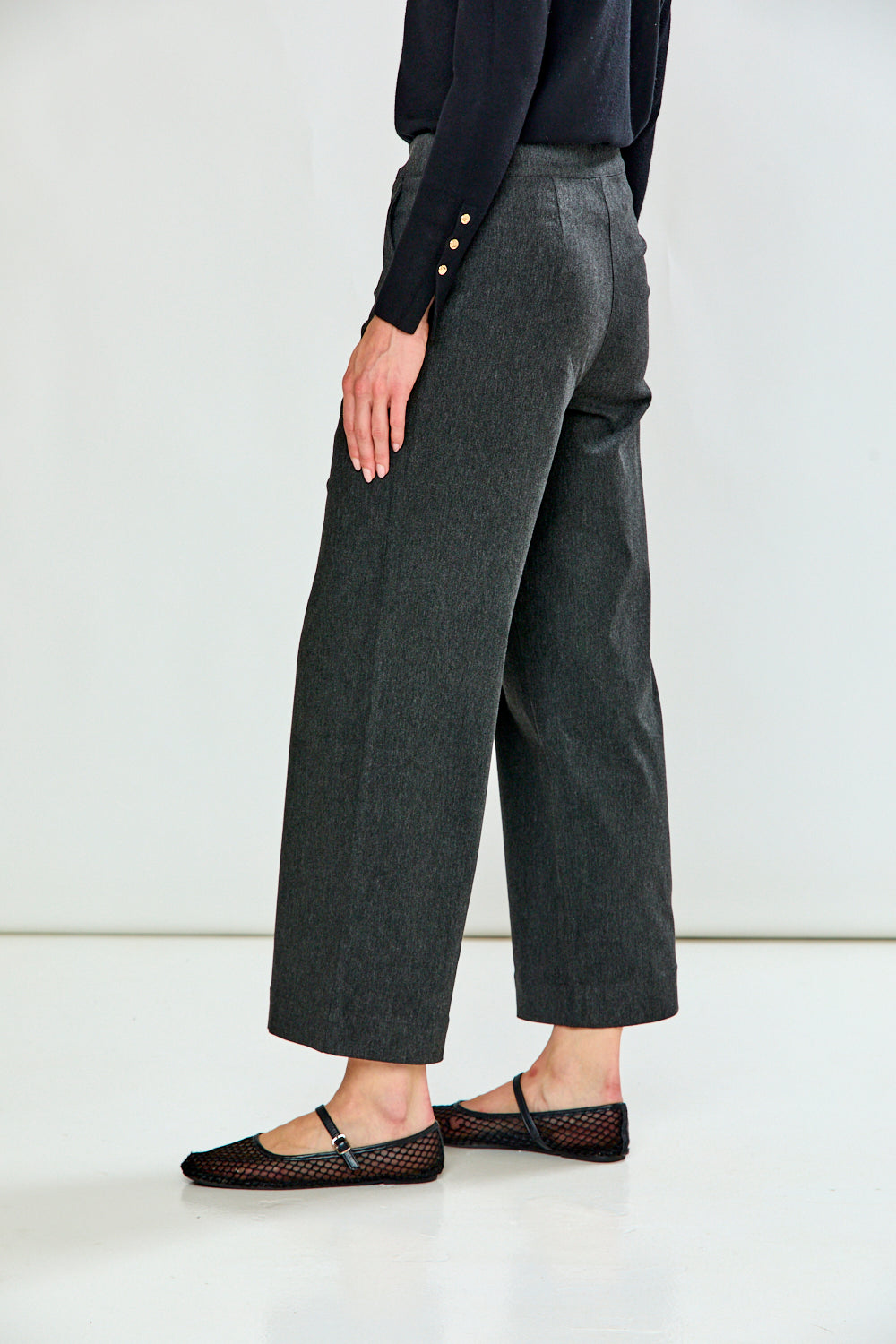 A person shown from the waist down wearing dark charcoal gray wide-leg cropped trousers, a black long-sleeve top, and black flat shoes with a fishnet pattern, set against a plain white studio background.