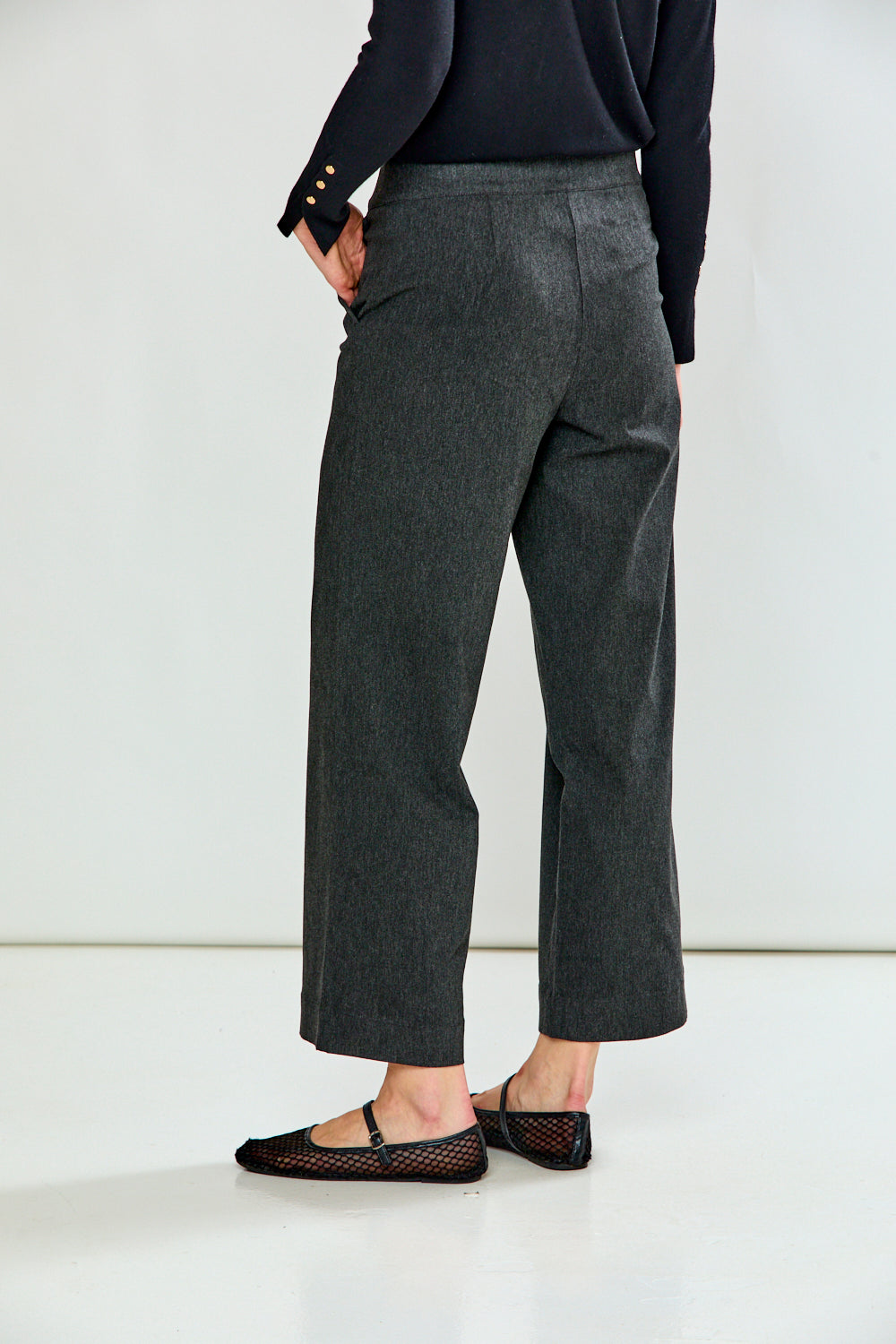 A person shown from the back wearing dark charcoal gray wide-leg cropped trousers, a black long-sleeve top, and black flat shoes against a clean white studio background.