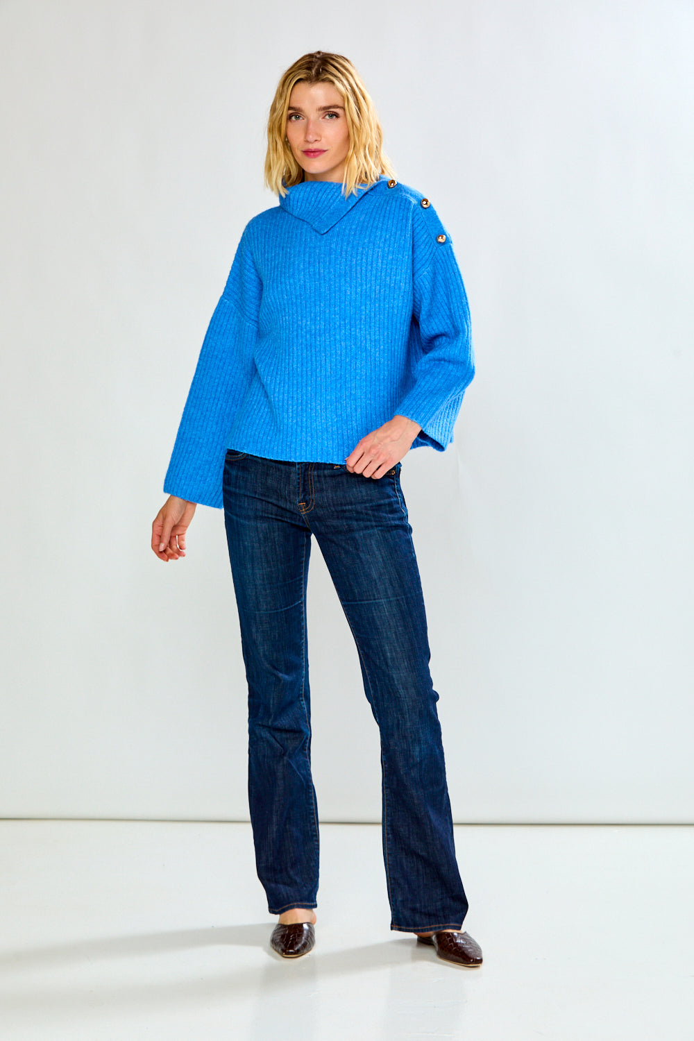 A woman modeling a bright blue ribbed knit sweater with a high mock neck and decorative buttons on the shoulders, paired with dark blue jeans, against a light gray studio background.