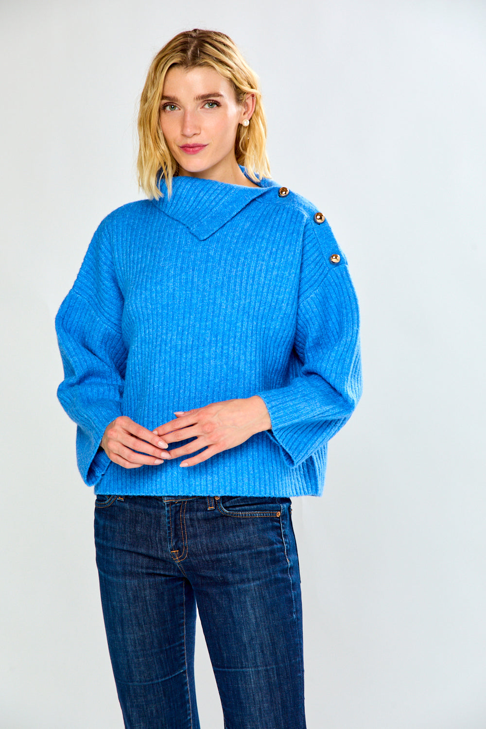 A woman modeling a bright blue ribbed knit sweater with a high mock neck and decorative buttons on the shoulders, paired with dark blue jeans, against a light gray studio background.