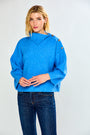 A woman modeling a bright blue ribbed knit sweater with a high mock neck and decorative buttons on the shoulders, paired with dark blue jeans, against a light gray studio background.