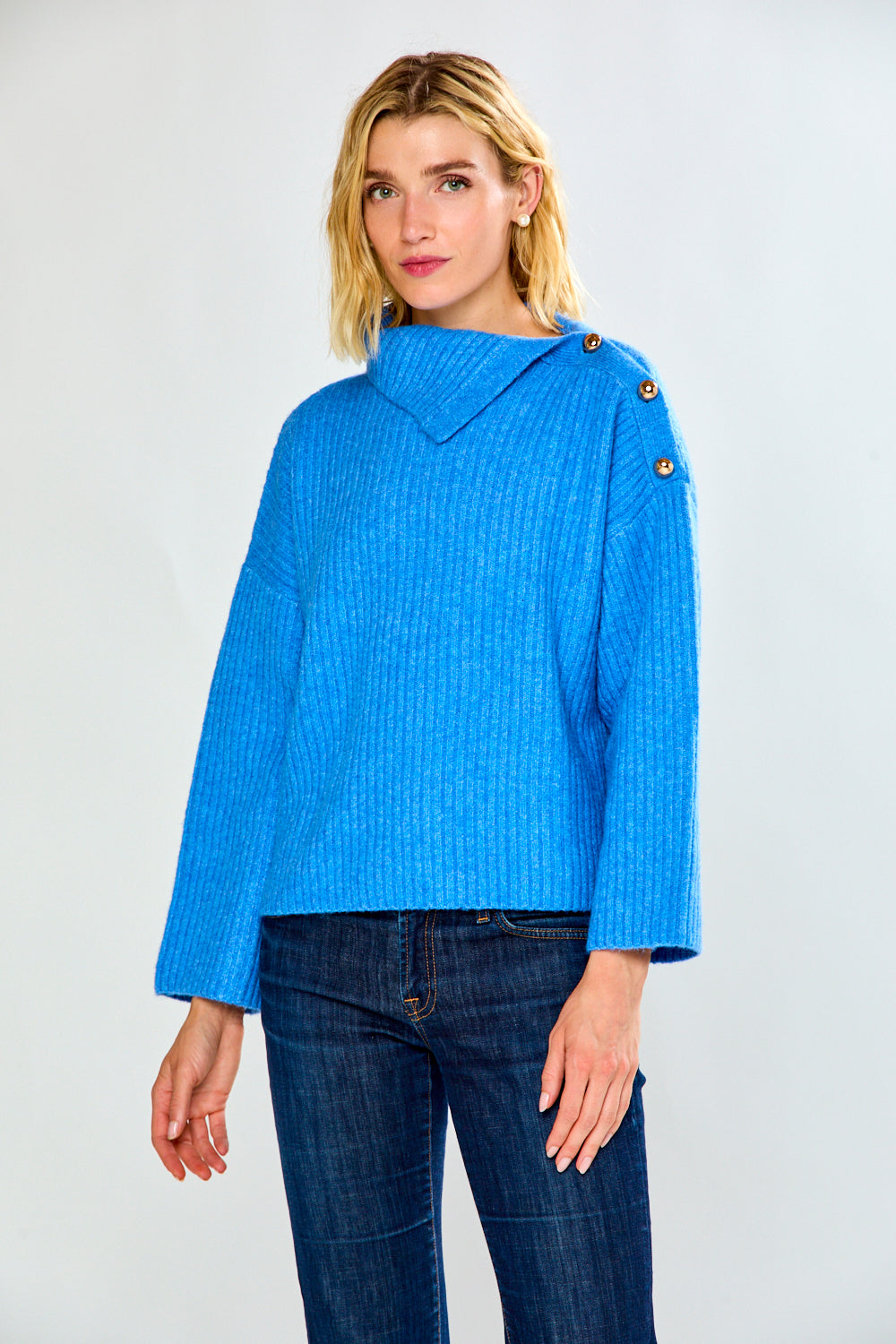 A woman modeling a bright blue ribbed knit sweater with a high mock neck and decorative buttons on the shoulders, paired with dark blue jeans, against a light gray studio background.