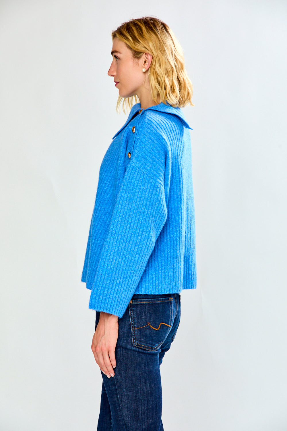 A woman modeling a bright blue ribbed knit sweater with a high mock neck and decorative buttons on the shoulders, paired with dark blue jeans, against a light gray studio background.