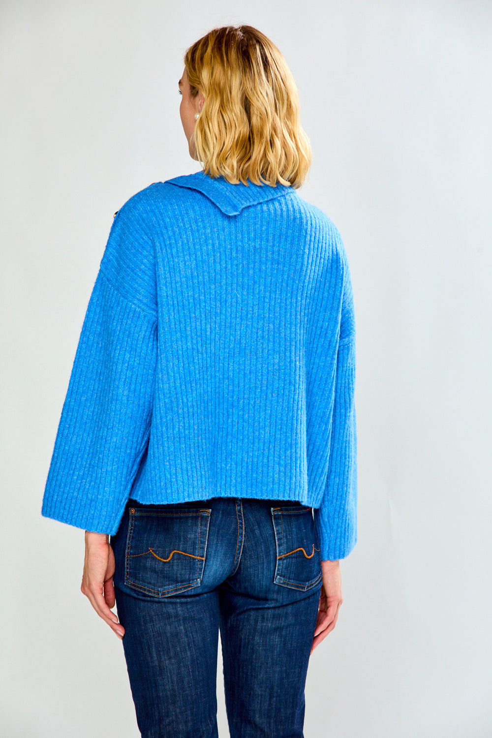 A woman modeling a bright blue ribbed knit sweater with a high mock neck and decorative buttons on the shoulders, paired with dark blue jeans, against a light gray studio background.