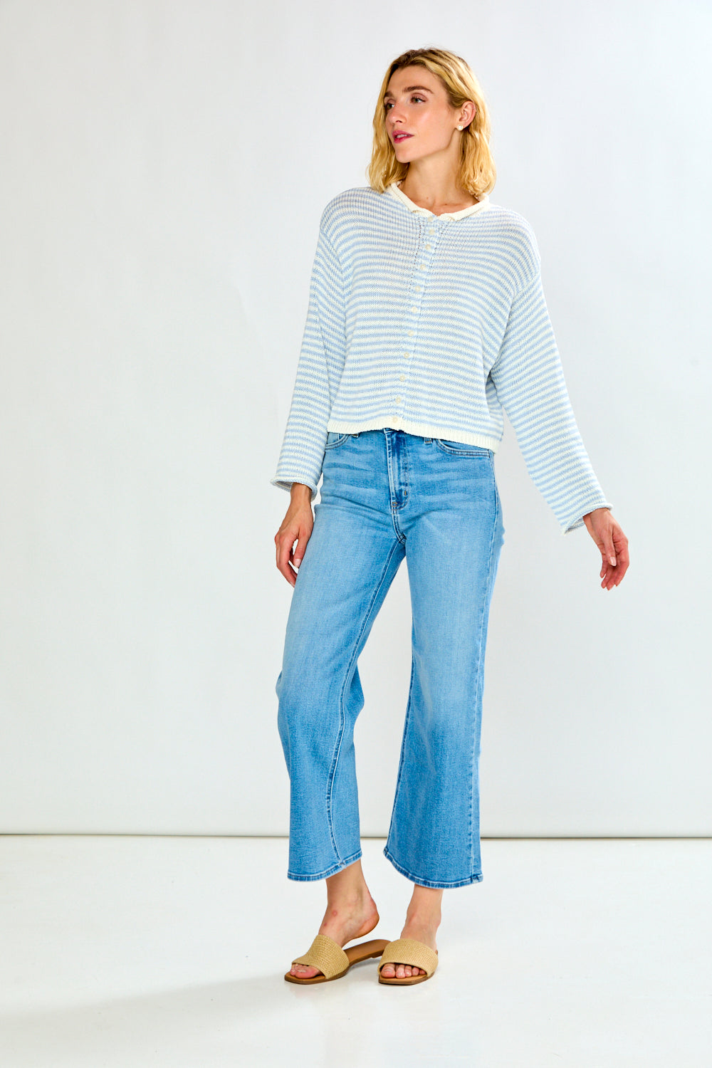 A woman modeling a light blue and white striped long-sleeve sweater with a button-up front, paired with light-wash blue jeans, against a plain light gray studio background.