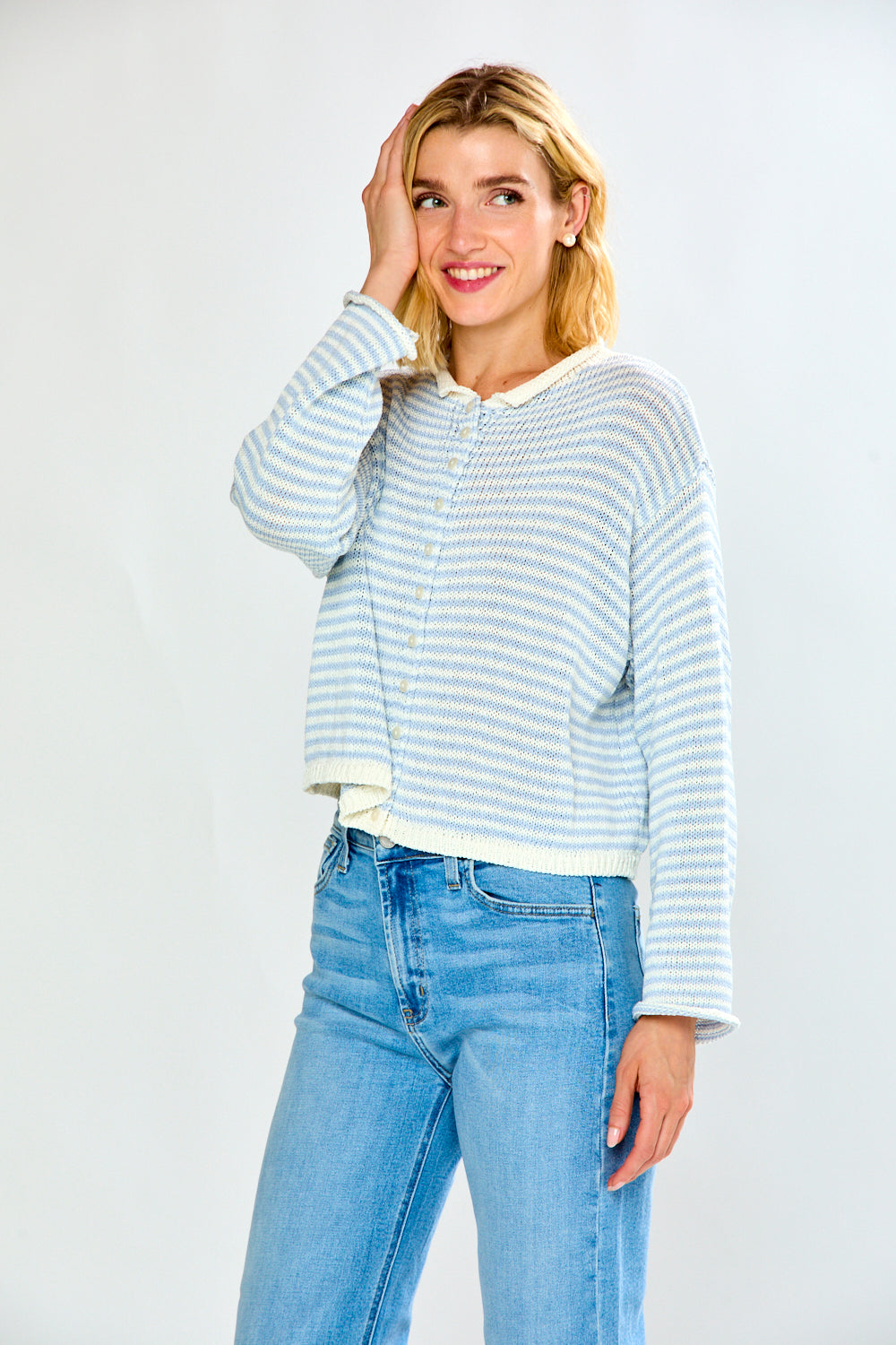 A woman modeling a light blue and white striped cropped knit sweater with a cream collar and button placket, paired with light-wash blue jeans, against a plain light gray studio background.