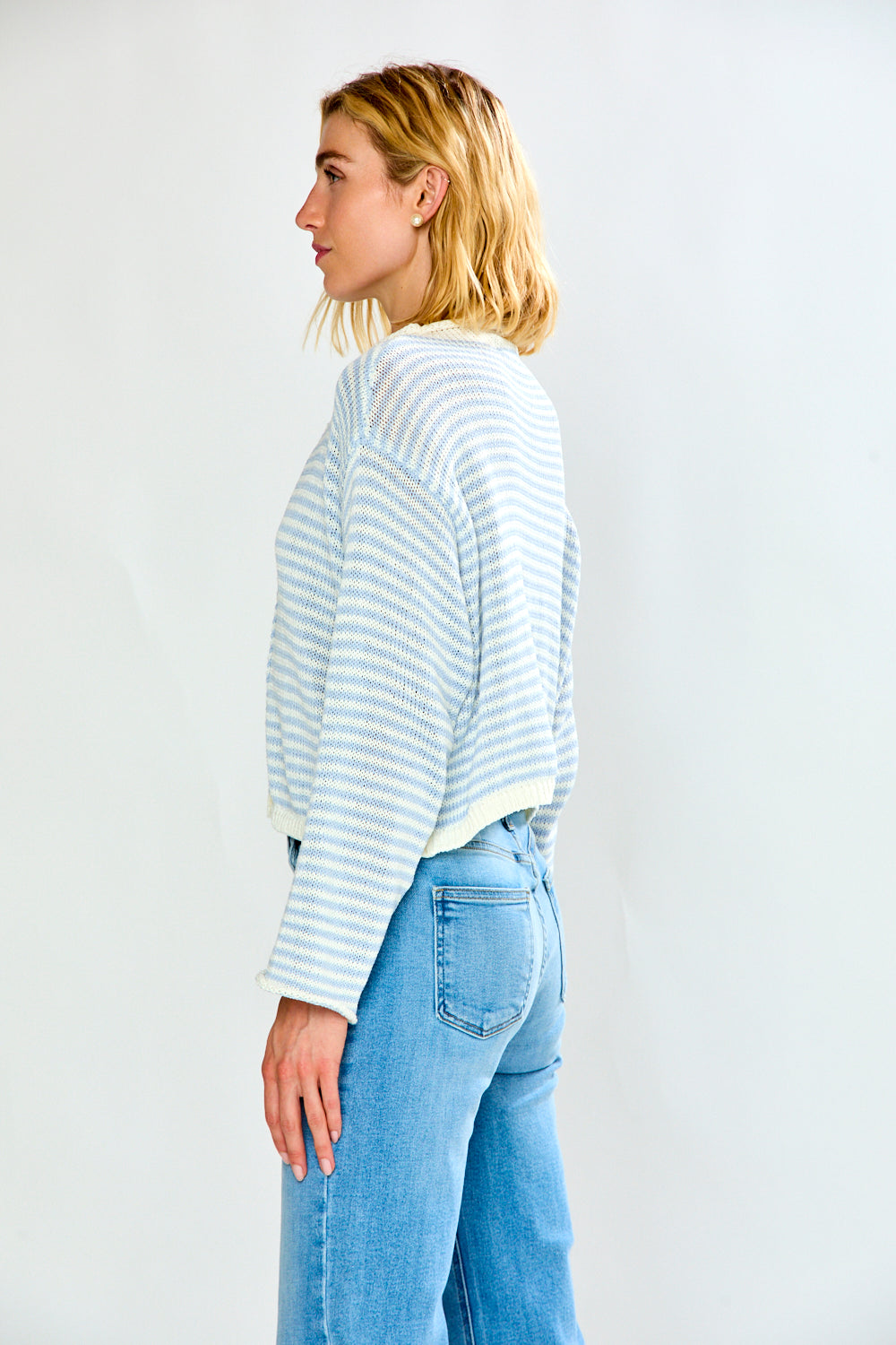 A woman modeling a light blue and white striped long-sleeve sweater with a button-up front, paired with light-wash blue jeans, against a plain light gray studio background.