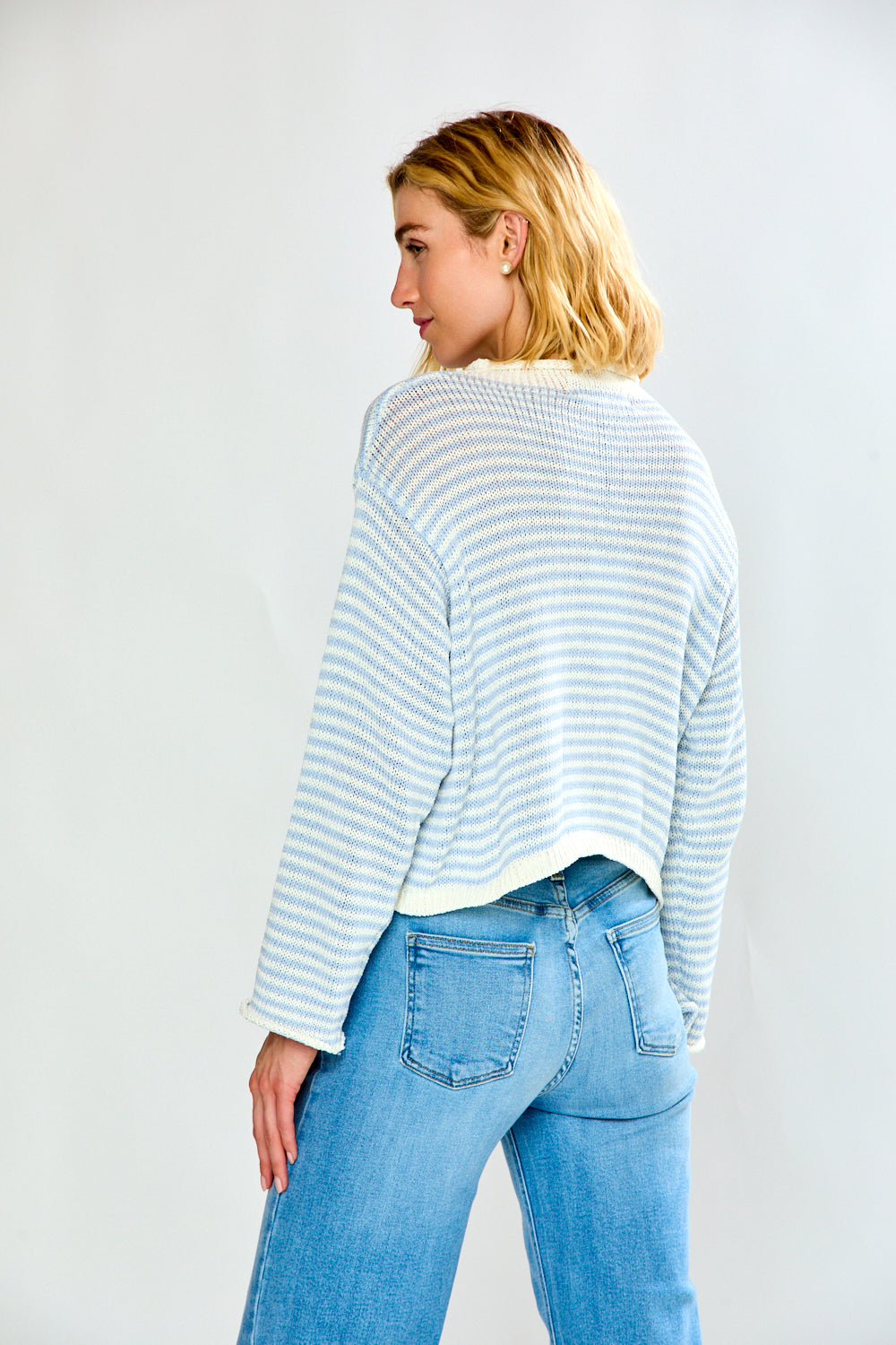 A woman modeling a light blue and white striped long-sleeve sweater with a button-up front, paired with light-wash blue jeans, against a plain light gray studio background.