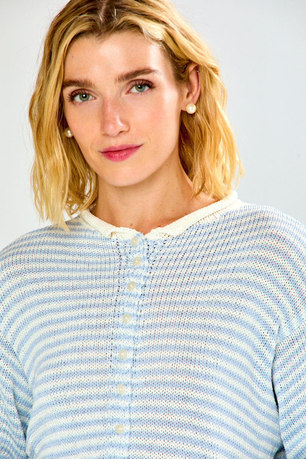A woman modeling a light blue and white striped long-sleeve sweater with a button-up front, paired with light-wash blue jeans, against a plain light gray studio background.