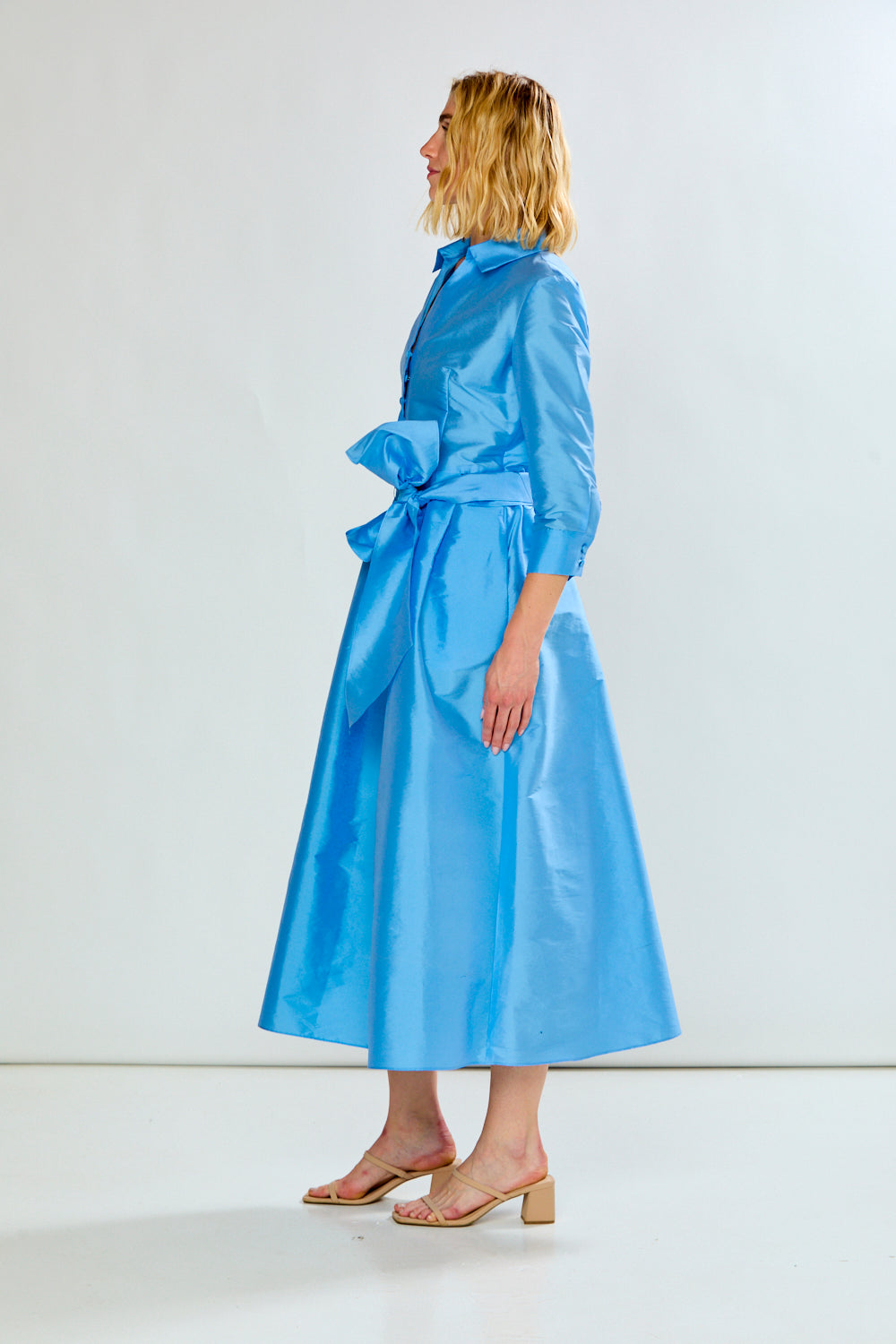The Ainsley Dress - Light Blue shown in side view with a collared neckline long sleeves tied waist and flowing midi skirt for a sophisticated event look