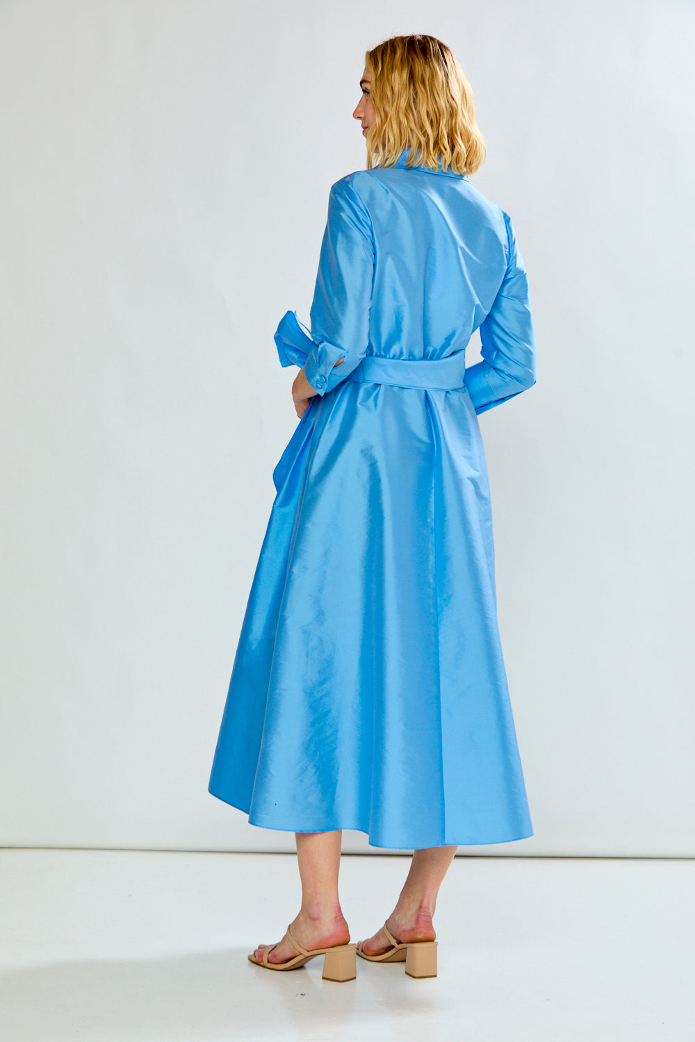 The Ainsley Dress - Light Blue shown from the back on a model with long sleeves a tied waist a flowing midi skirt and heeled sandals