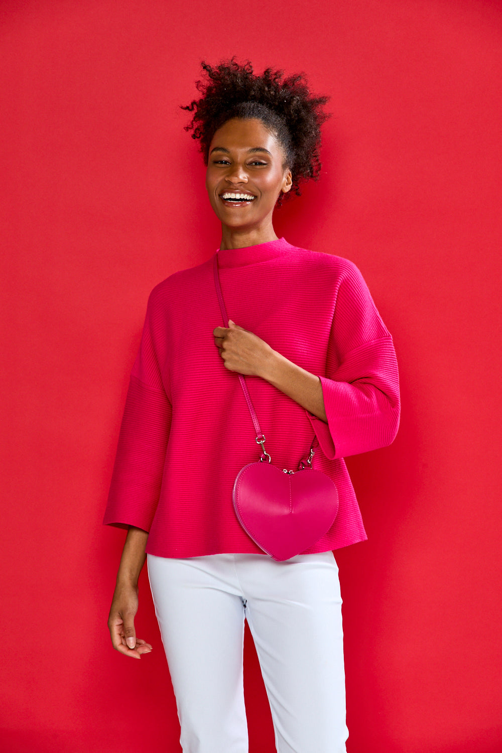 Woman in a bright pink top and white pants holding a matching pink handbag against a red background