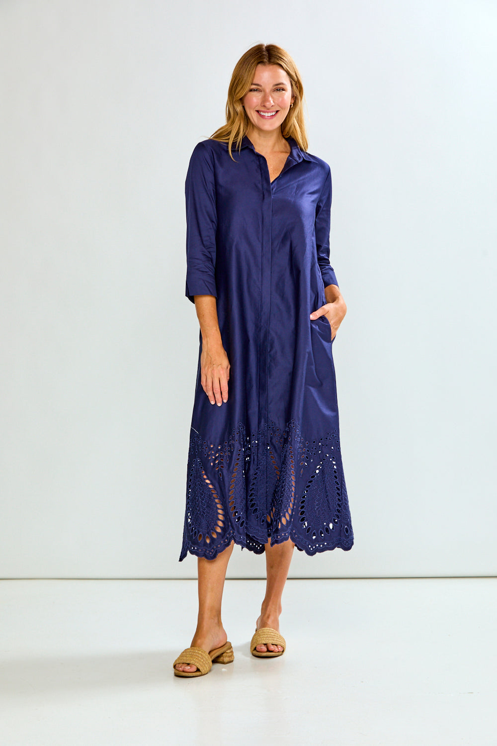 A woman modeling a navy blue midi shirt dress with three-quarter sleeves, a button-down front, and eyelet lace detailing along the hem, paired with tan slide sandals against a plain white studio background.