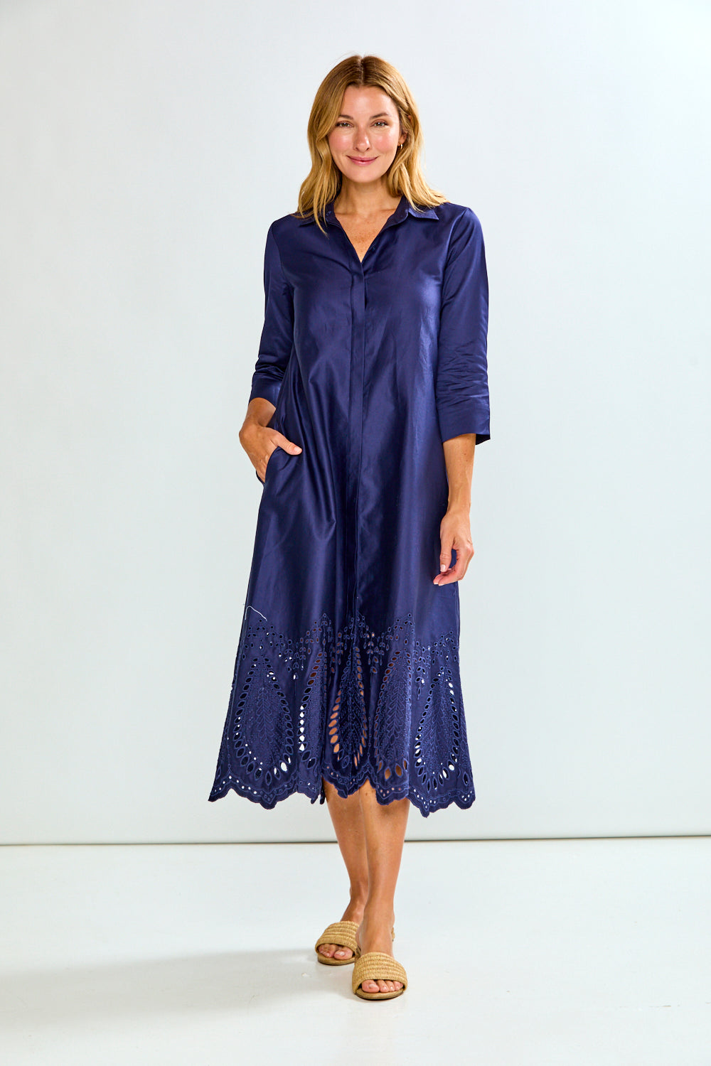 A woman modeling a navy blue midi shirt dress with three-quarter sleeves, a button-down front, and eyelet lace detailing along the hem, paired with tan slide sandals against a plain white studio background.