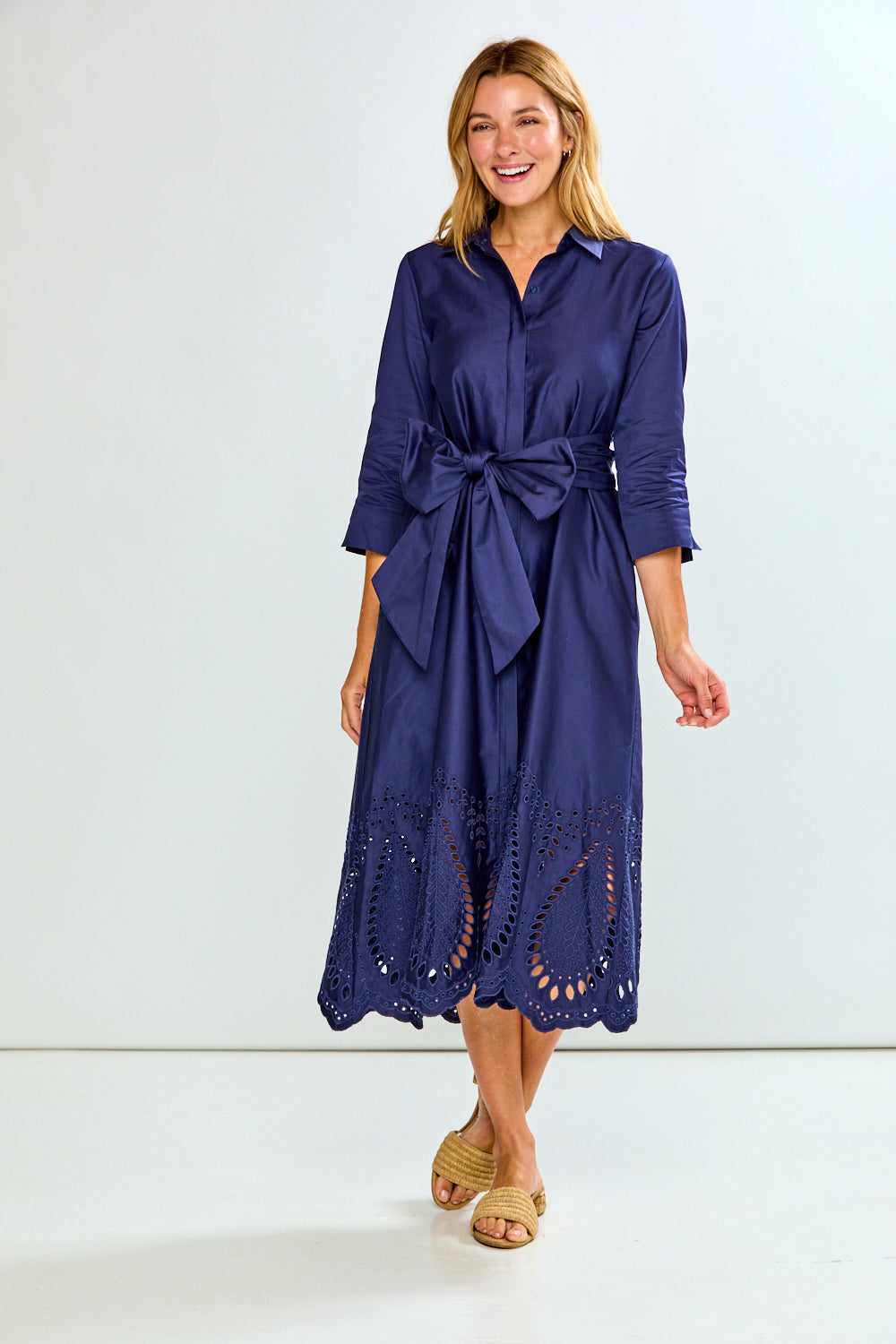 A woman modeling a navy blue midi shirt dress with three-quarter sleeves, a button-down front, and eyelet lace detailing along the hem, paired with tan slide sandals against a plain white studio background.