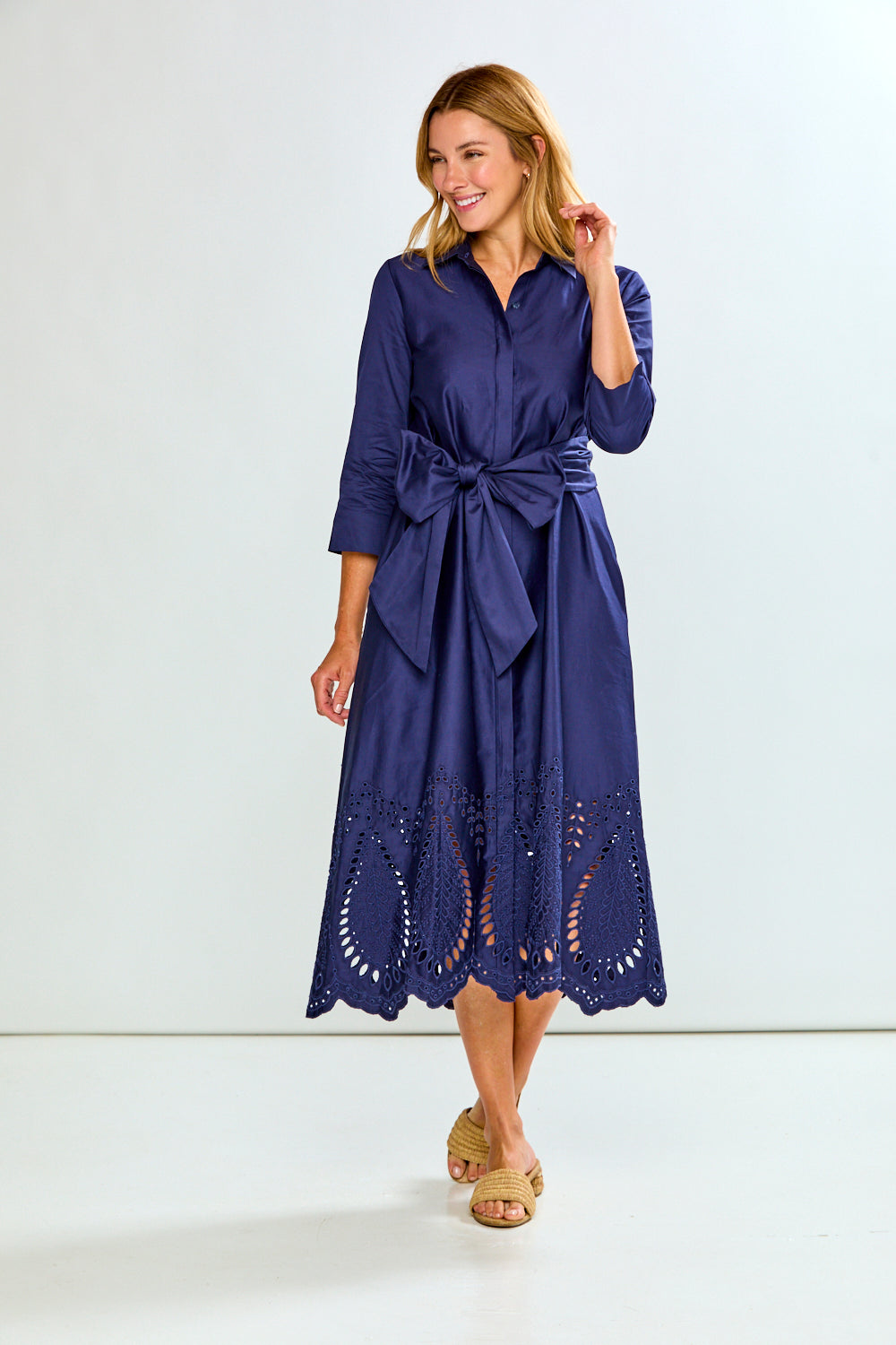 A woman modeling a navy blue midi shirt dress with three-quarter sleeves, a button-down front, and eyelet lace detailing along the hem, paired with tan slide sandals against a plain white studio background.
