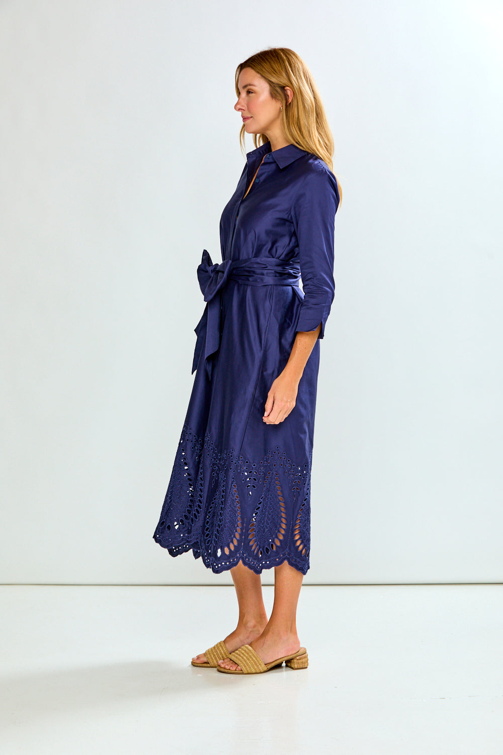 A woman modeling a navy blue midi shirt dress with three-quarter sleeves, a button-down front, and eyelet lace detailing along the hem, paired with tan slide sandals against a plain white studio background.