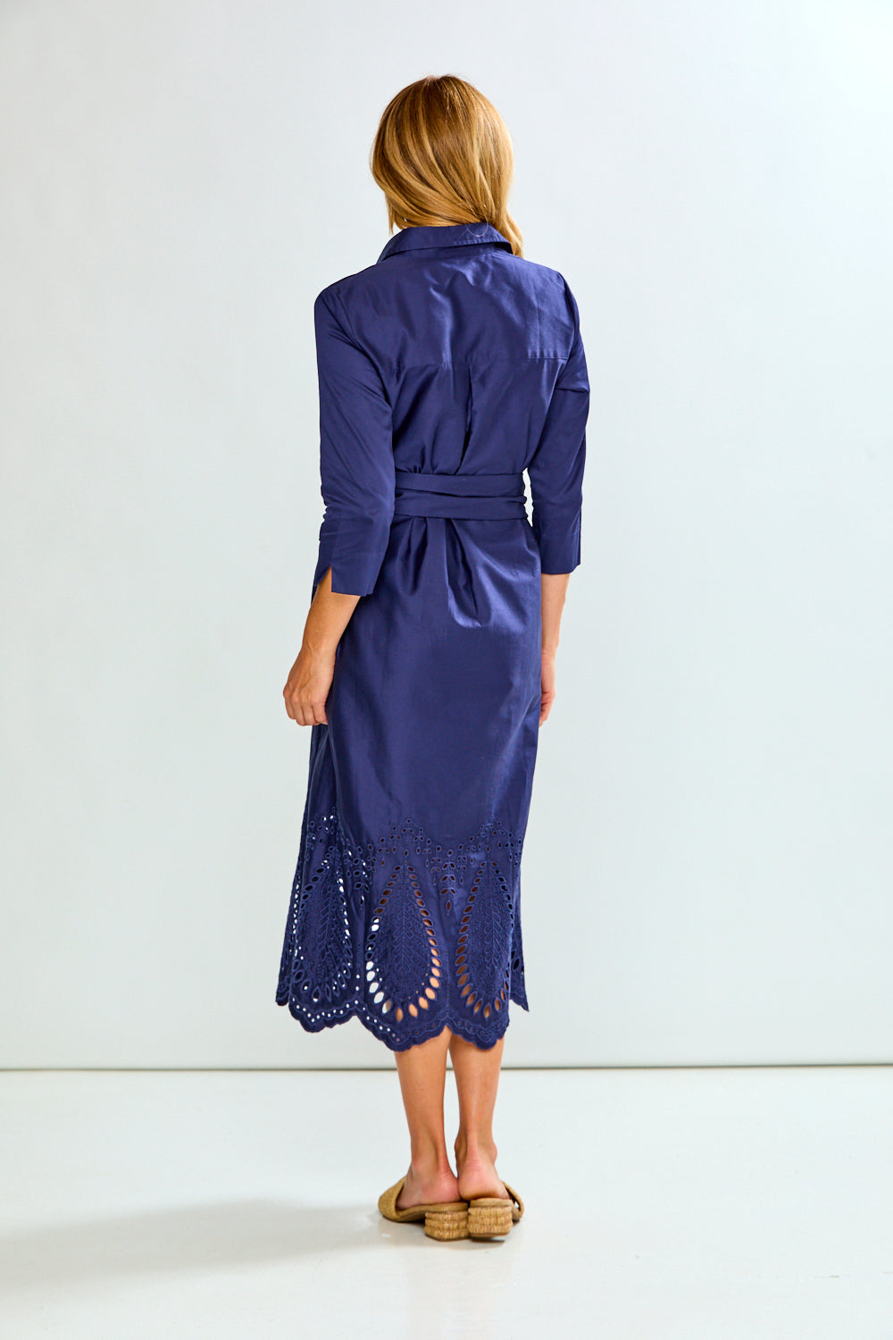 A woman modeling a navy blue midi shirt dress with three-quarter sleeves, a button-down front, and eyelet lace detailing along the hem, paired with tan slide sandals against a plain white studio background.
