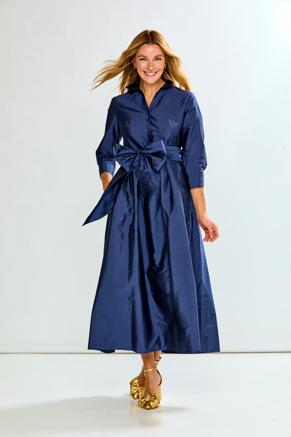 A woman modeling a navy blue midi shirt dress with a matching waist tie, paired with gold platform sandals, against a plain white studio background.
