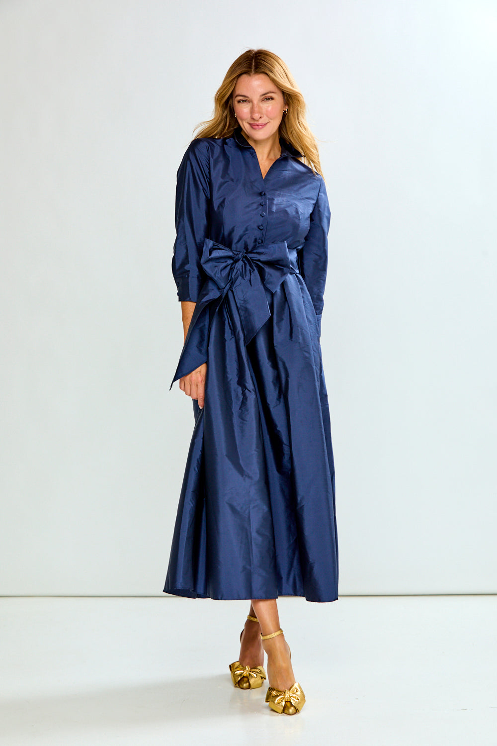 A woman modeling a navy blue midi shirt dress with three-quarter sleeves, a button-up front, and a matching fabric tie belt at the waist, paired with gold heels against a plain white studio background.