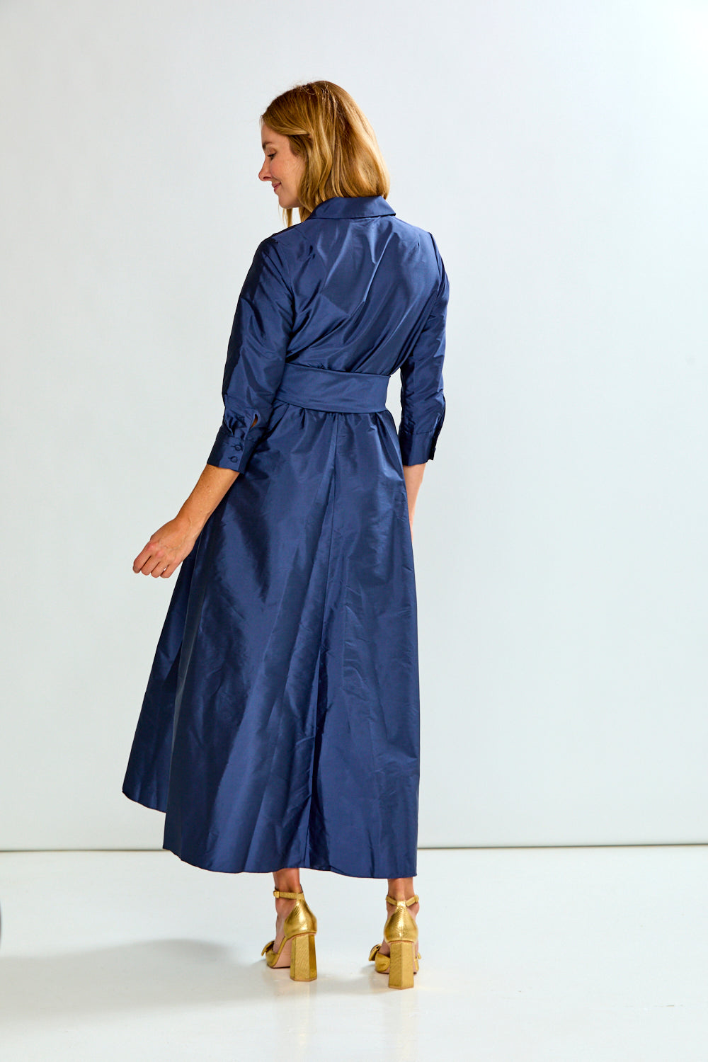 A woman wearing a navy blue satin midi dress with a waist tie, shown from the back against a plain white studio background.