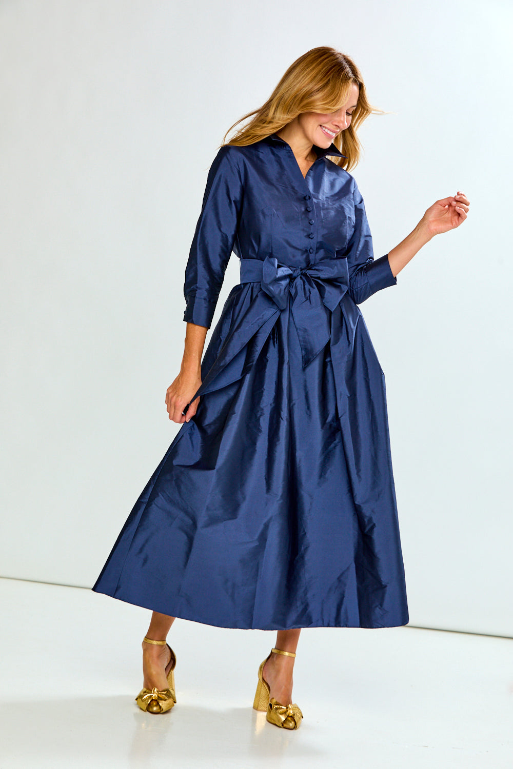 A woman modeling a navy blue midi shirt dress with three-quarter sleeves, a button-up front, and a matching self-tie belt at the waist, paired with gold high heels against a plain white studio background.