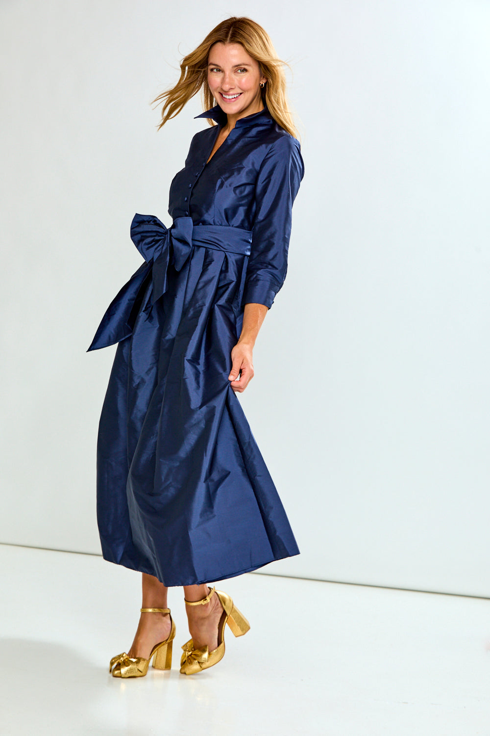 A woman modeling a navy blue satin midi shirt dress with a matching waist tie, paired with metallic gold high-heeled sandals, against a plain white studio background.